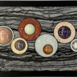 Abyssal Origins <br>
9" x 15.625" x 2.5", 2024 (September) <br>
Marble, limestone, cuprite with chrysocolla; spheres of silicified porcelanite and aventurescent feldspar, ruby with in crystal, jadeite, banded agate, chevron amethyst, and orbicular jasper. A group of coins sitting on top of a wooden table.