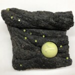 Jewel of the Pacific <br>
7 1/4" x 6 1/2," 2019 <br>
Basalt, serpentinite sphere, faceted peridot A black bag with green beads on it