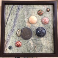Earth Sampler Series XVI: Black Ammonite with Cubic Pyrite <br> 13” x 13”, 2016 <br> Serpetinite; ammonite, pyrite; spheres of banded jasper, unakite, sunstone, peach calcite, indigo gabbro, snakeskin jasper, orbicular jasper A picture of some shells and rocks in a frame.