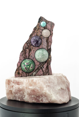 Sculpture VI: Earth Murmurs <br>
12.5” x 9.5” x 8.5”, 2023 <br>
Rose quartz base; Front: lepidolite pegmatite; spheres of rhodochrosite, banded agate, snowflake obsidian, rhodonite, calcite; Back: spiderweb rhodonite; spheres of larimar, rose quartz, amethyst, unknown green and white sphere, ruby and fuchsite A rock with several different colored stones on top of it.