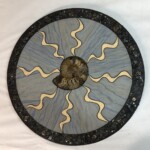 Circle of Spirals Revisited (II) <br> 23” diameter, 2021 <br> Fossil ammonite, dumortierite quartzite, limestone, syenite, pyritized ammonites A circular wooden plaque with a design of sun and waves.